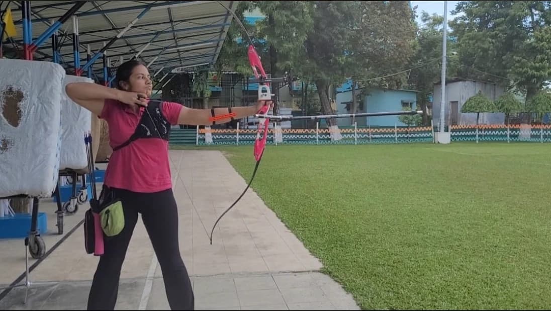 At full draw with a recurve bow on an outdoor range, target butts at the left of frame.